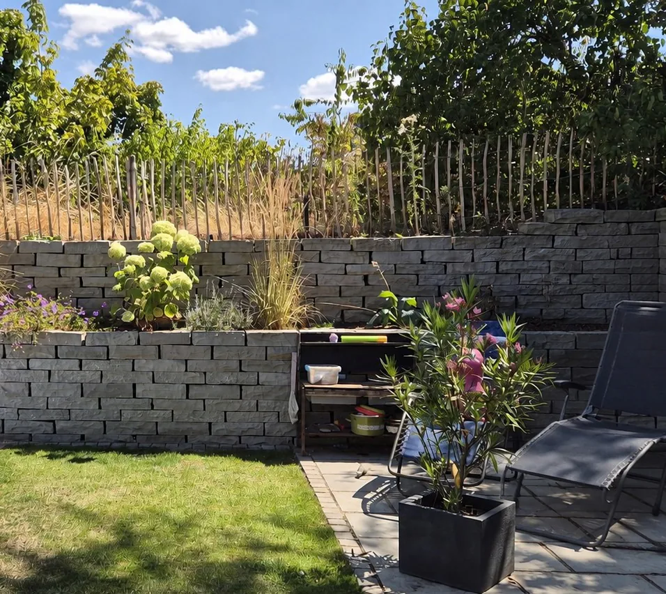 Zweistufige Natursteinmauer mit Bepflanzung, davor Rasen und eine gepflasterte Terrasse