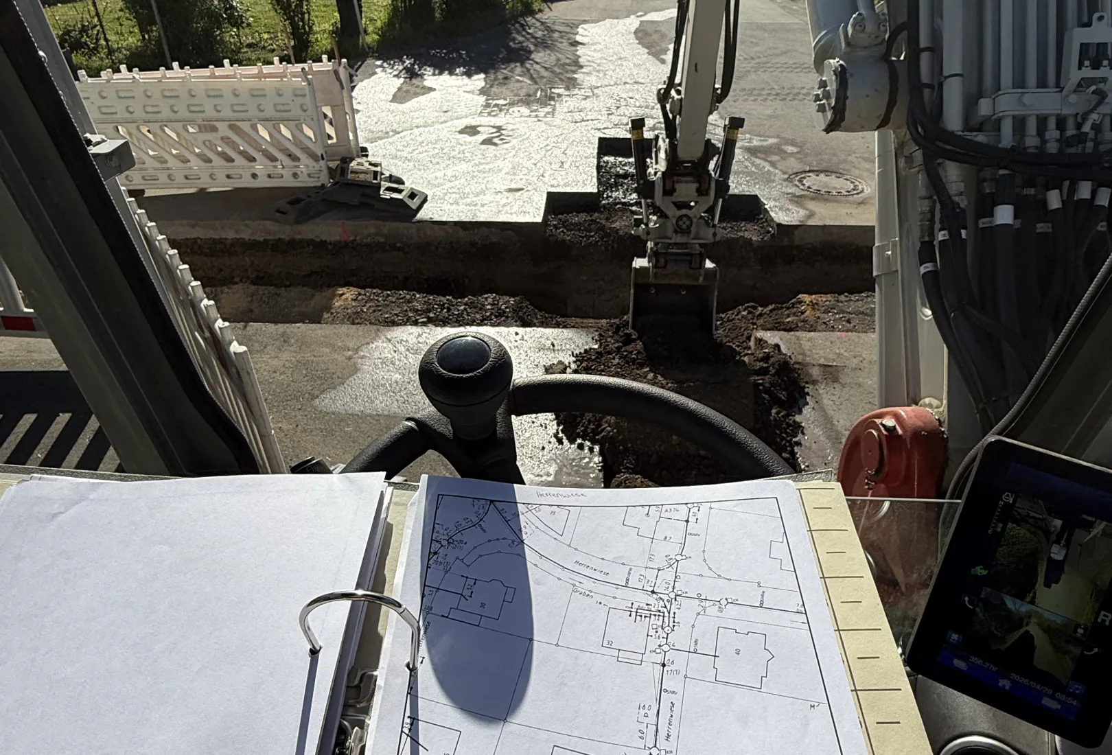 Blick aus der Baggerkabine über einen aufgeschlagenen Leitungsplan auf einen offenen Graben in einer Wohnstraße