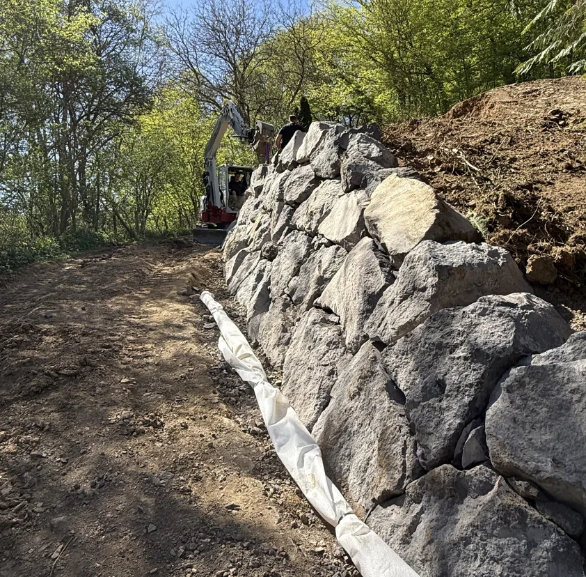 Neu gesetzte Natursteinmauer entlang eines Waldwegs, dahinter ein Minibagger von Valera