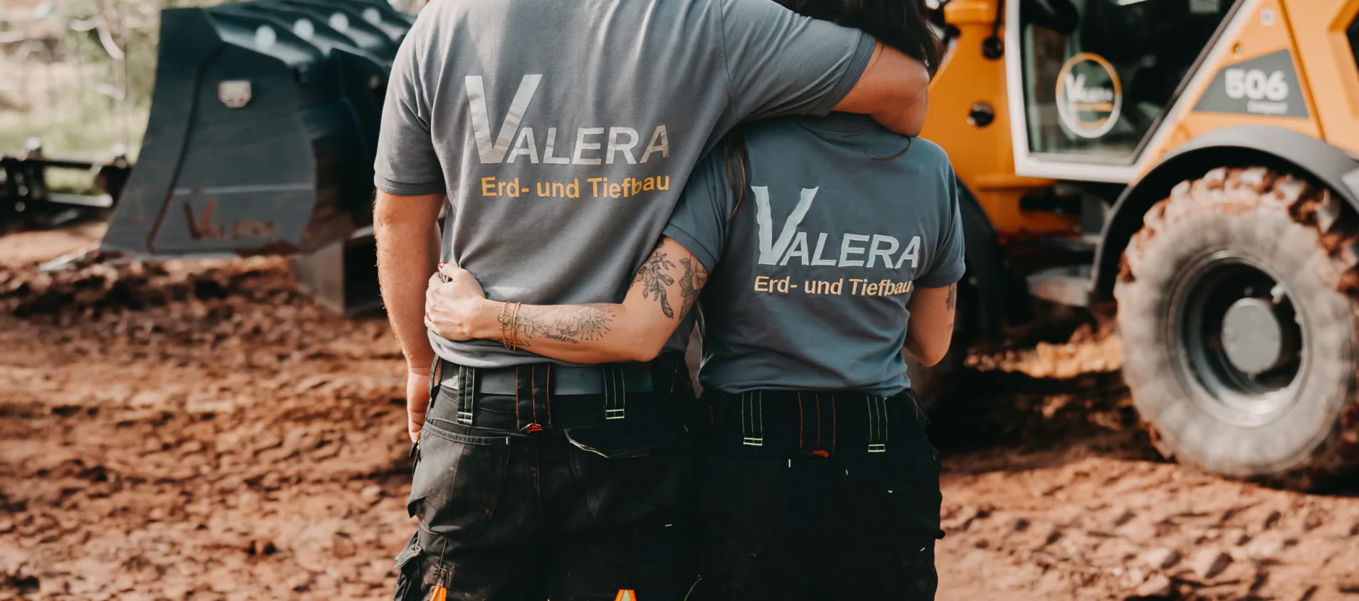 Zwei Mitarbeitende von Valera stehen Arm in Arm vor einem Mobilbagger und einem Radlader auf der Baustelle